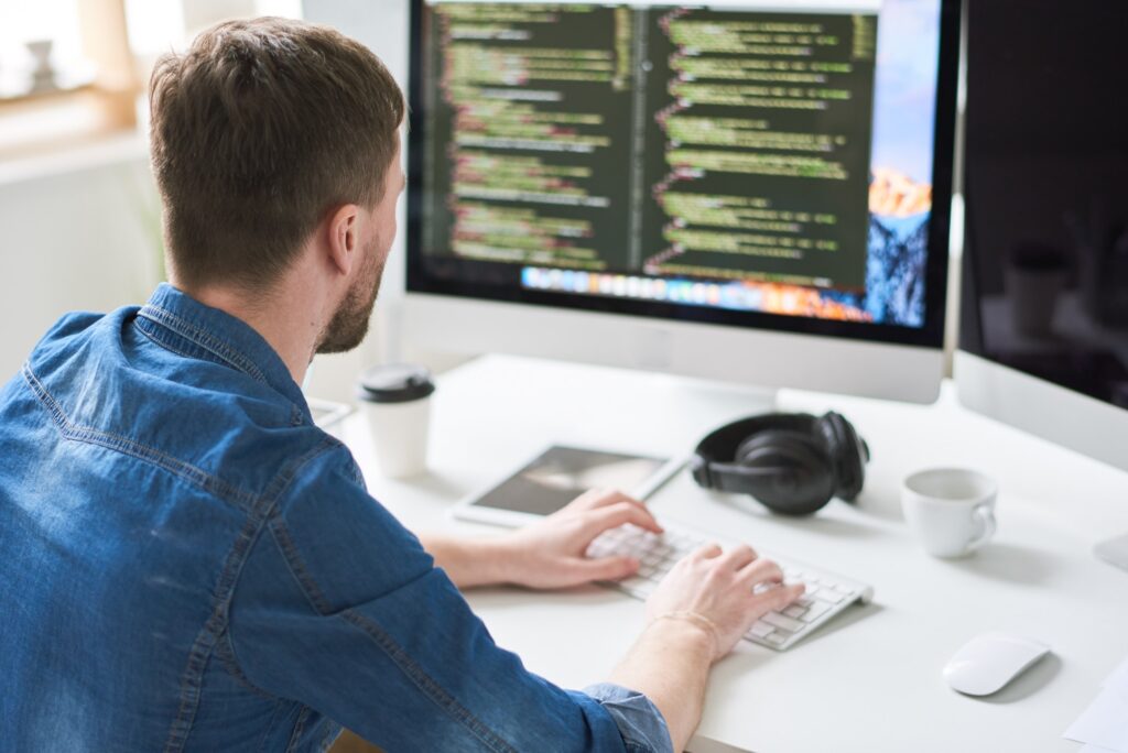A person in a denim shirt is sitting at a desk, typing on a keyboard in front of a large monitor displaying lines of code. A coffee cup, headphones, and mouse are also on the desk.
